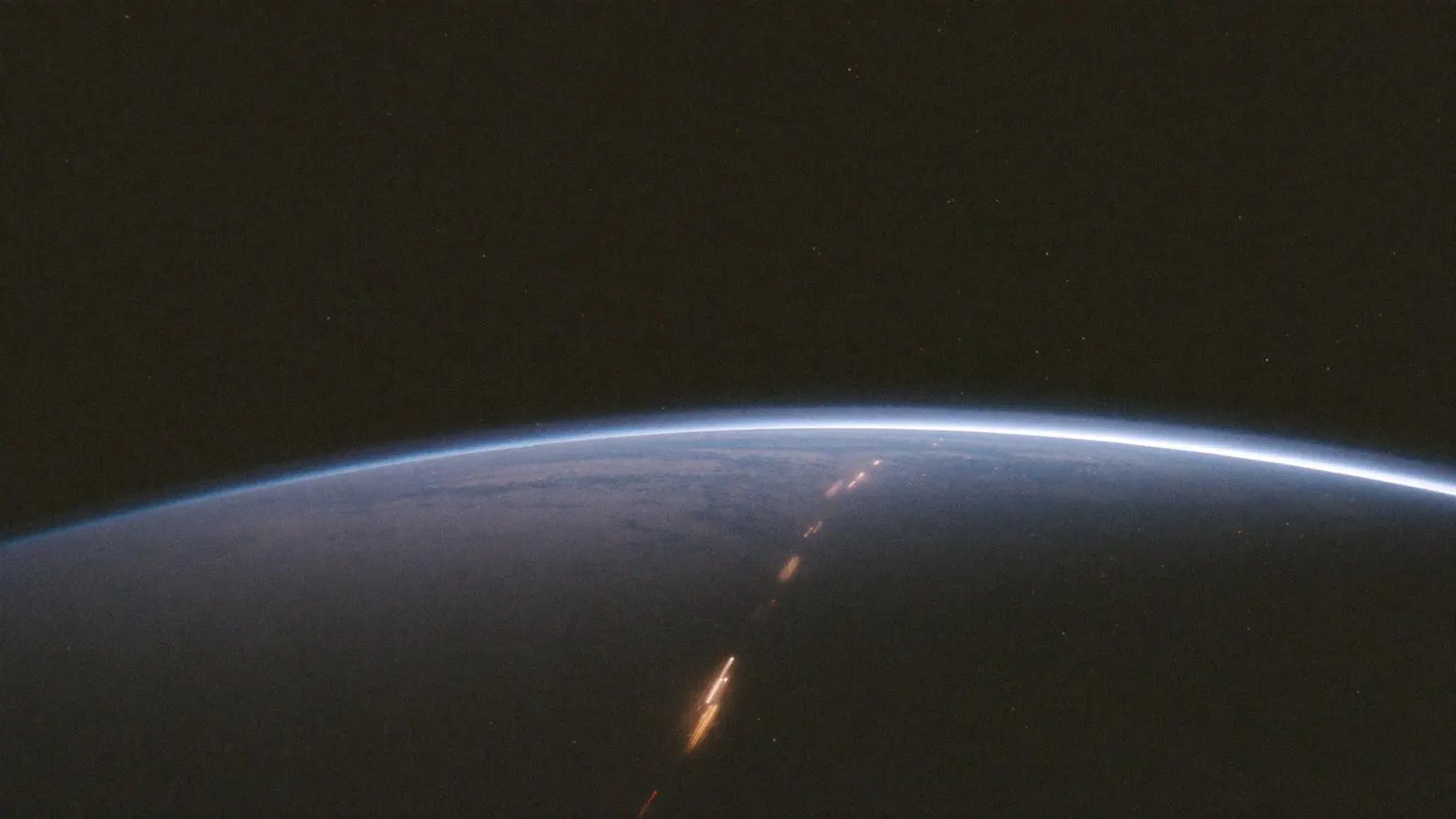 A settled world at night seen from orbit, its atmosphere a thin blue line and a transit corridor lit across its surface.